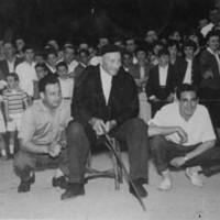 Keixeta aizkolaria plazan, aukian eserita, gizon biren artean. Atzean entzuleria --